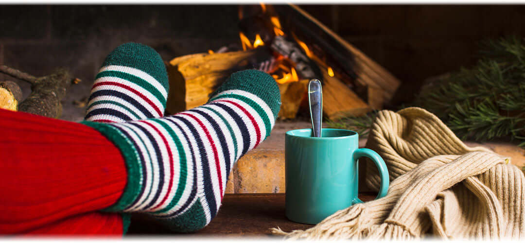 A person sitting by the fireplace, wrapped in a blanket, holding a hot drink, and wearing warm socks