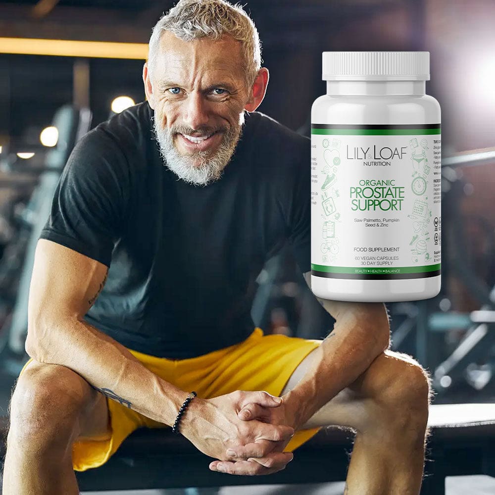 A smiling, fit middle-aged man with short grey hair and a trimmed beard, sitting and resting his arms on his knees. He's wearing a black t-shirt and yellow shorts. In the foreground, there's a product placement Organic Prostate Support