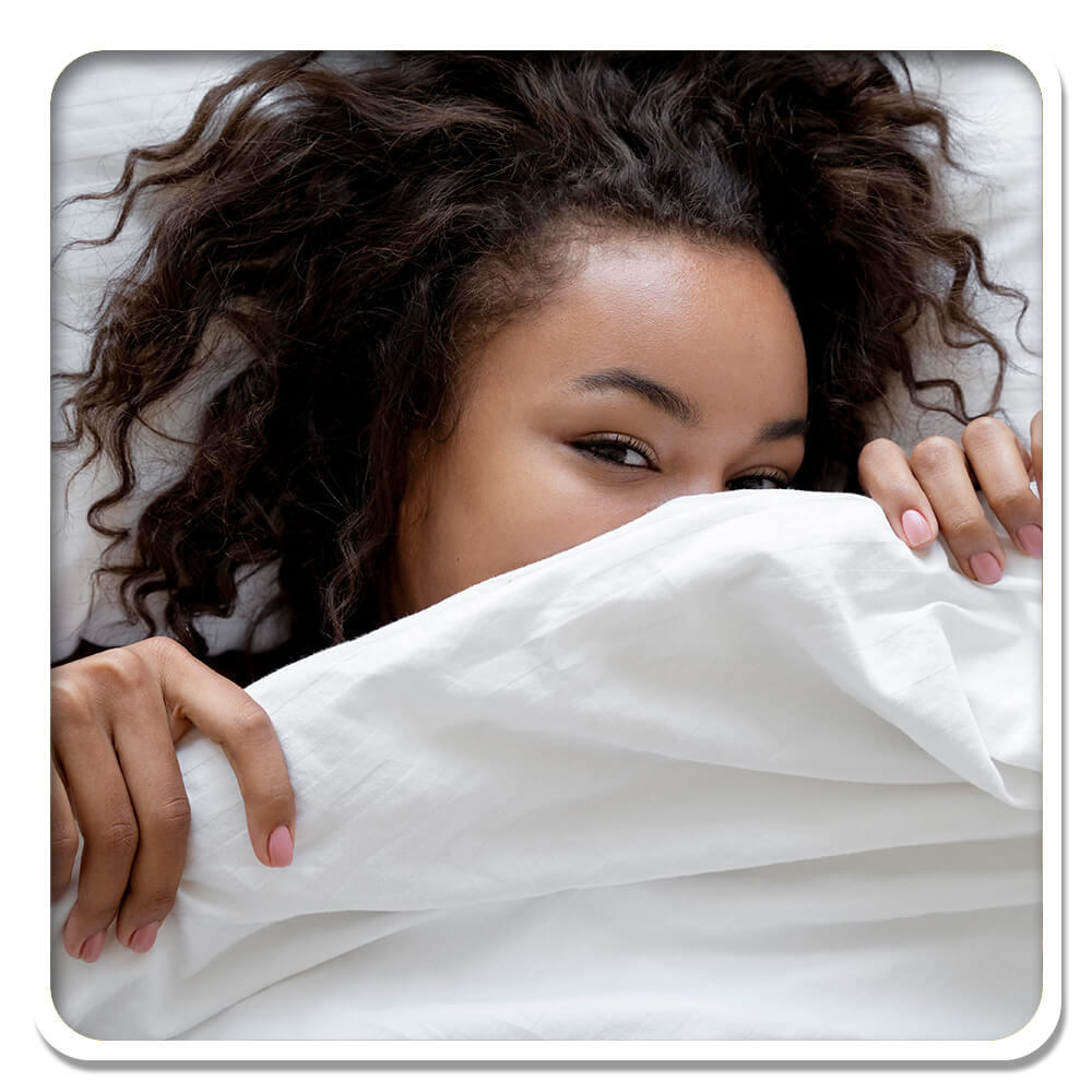A woman enjoys a restful sleep, waking up under a soft duvet with a radiant smile, refreshed from using Lily & Loaf's natural health supplements, ready to start her day with positivity and energy.