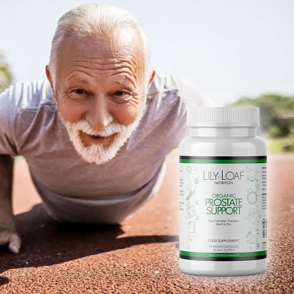 An elderly man with white hair and a white beard, smiling warmly at the camera while doing a push-up on a track. He is wearing a light grey t-shirt, and his fitness suggests an active and healthy lifestyle. Next to him is a prominently displayed bottle of Lily & Loaf's Organic Prostate Support dietary supplement